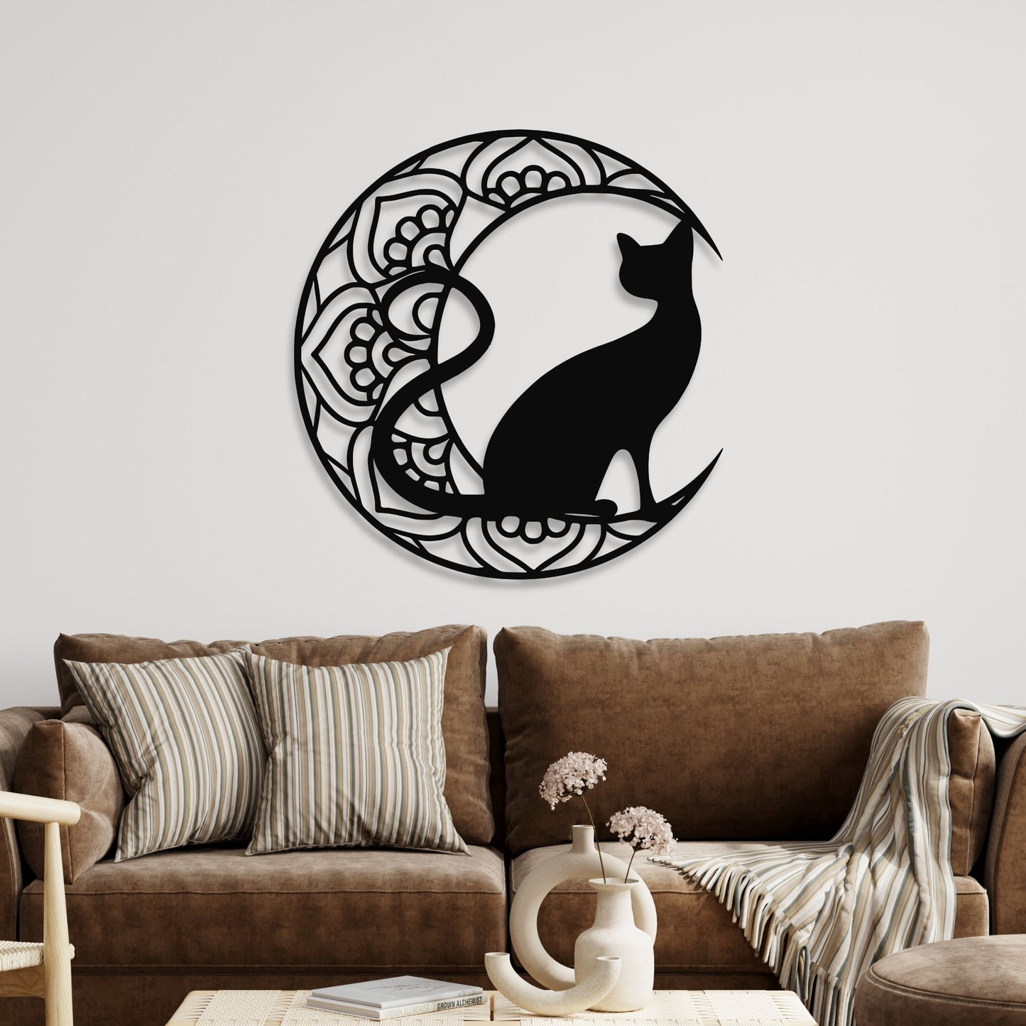 Decorative wall art of a black cat silhouette inside a circular mandala on a white wall above a brown sofa.