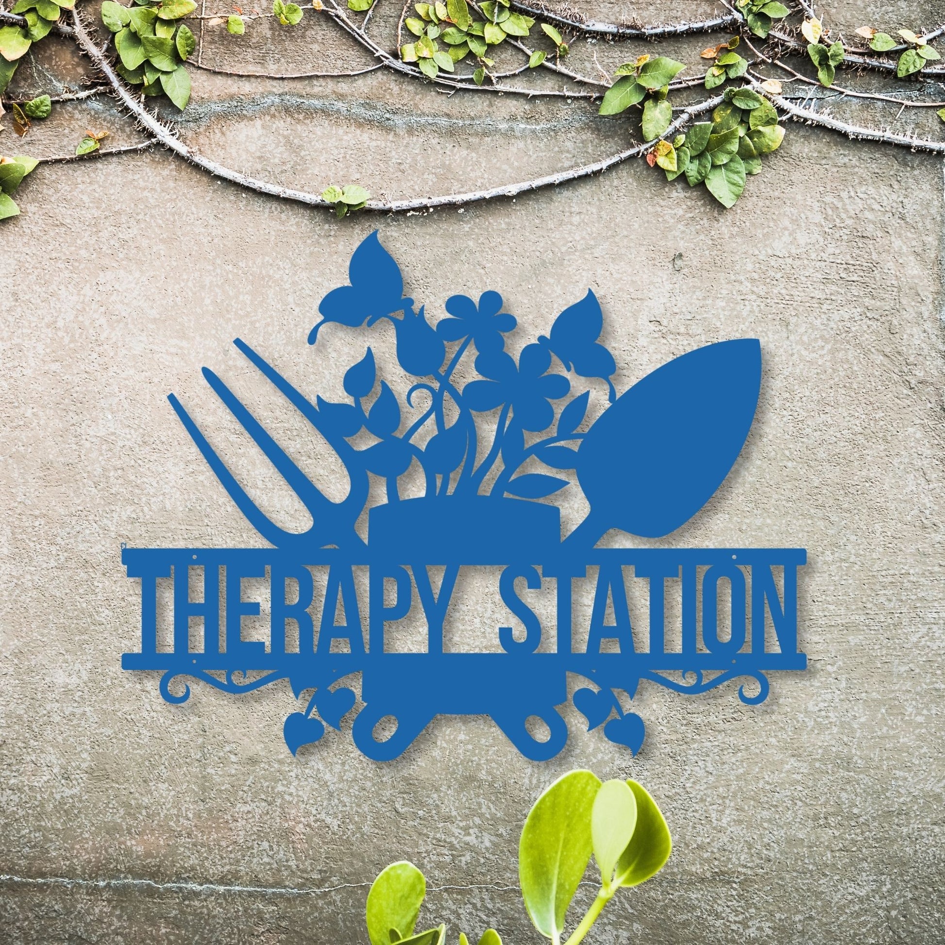 Blue metal sign with gardening tools and 'Therapy Station' text on a concrete surface with greenery.