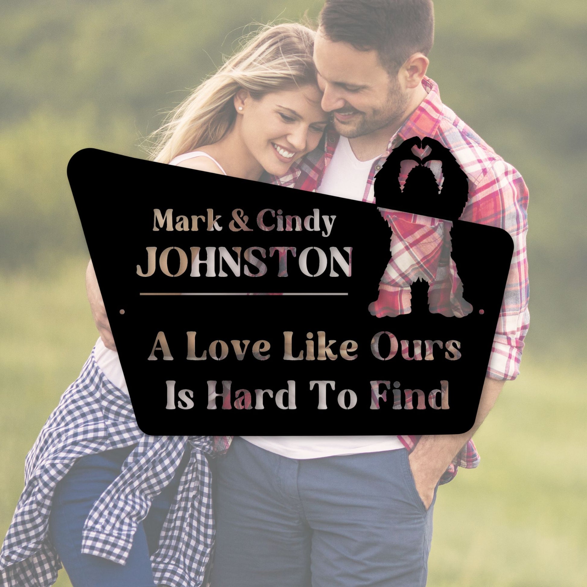 Couple embracing with a personalized sign between them, set against a blurred natural background.