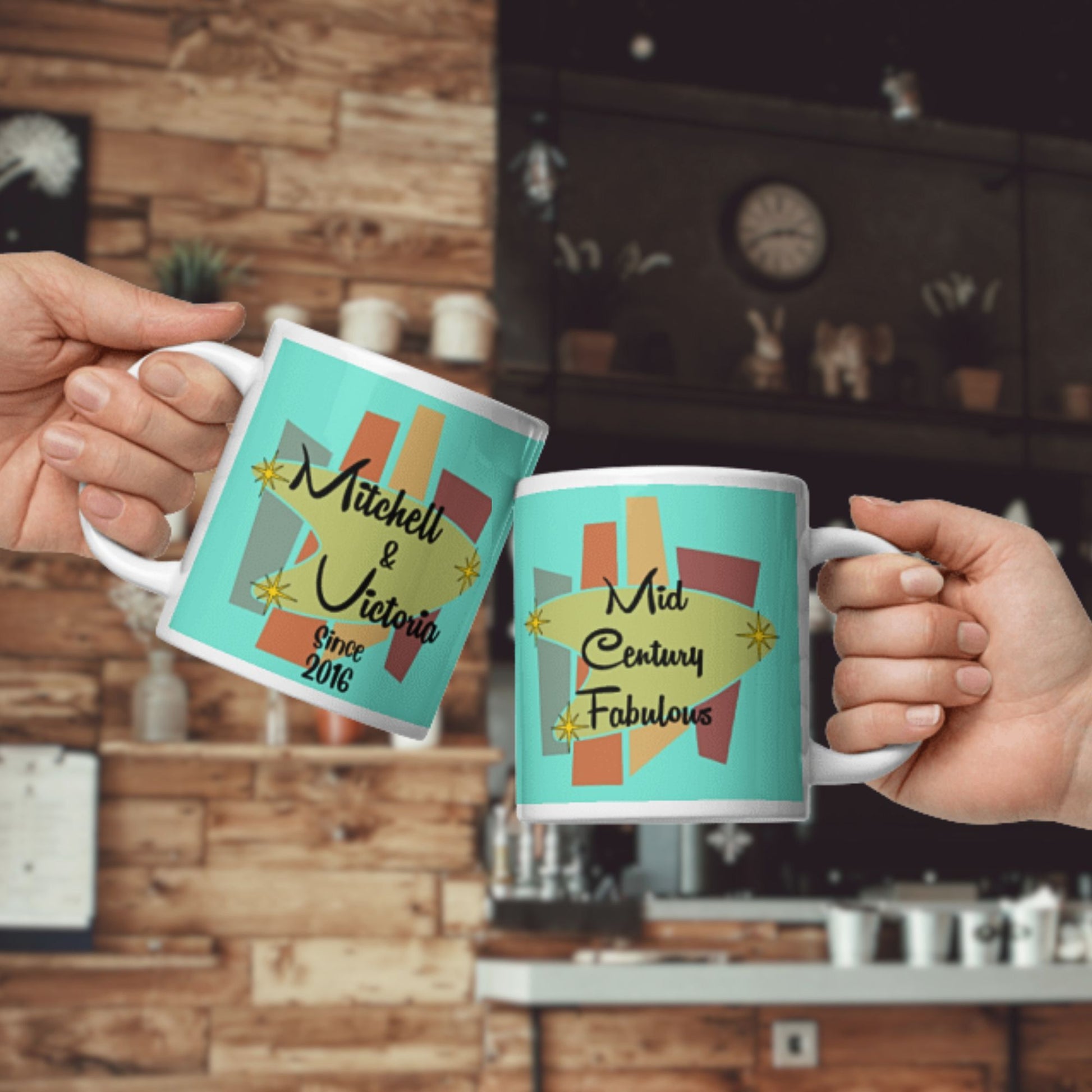 Two hands holding mugs with 'Mitchell & Victoria Since 2016' and 'Mid Century Fabulous' text in a kitchen setting.