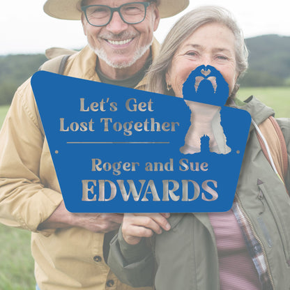 Two people holding a blue sign with text and a heart symbol, standing in a field.