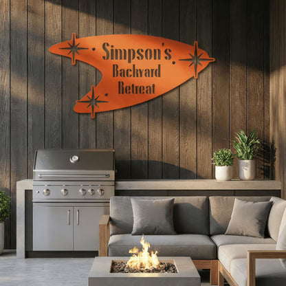 Outdoor living space with a grill, sofa, and fire pit against a wooden wall with a decorative sign.