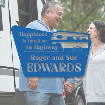 Two people holding hands with a blue sign behind them that reads 'Happiness is found on the Highway Roger and Sue Edwards'.