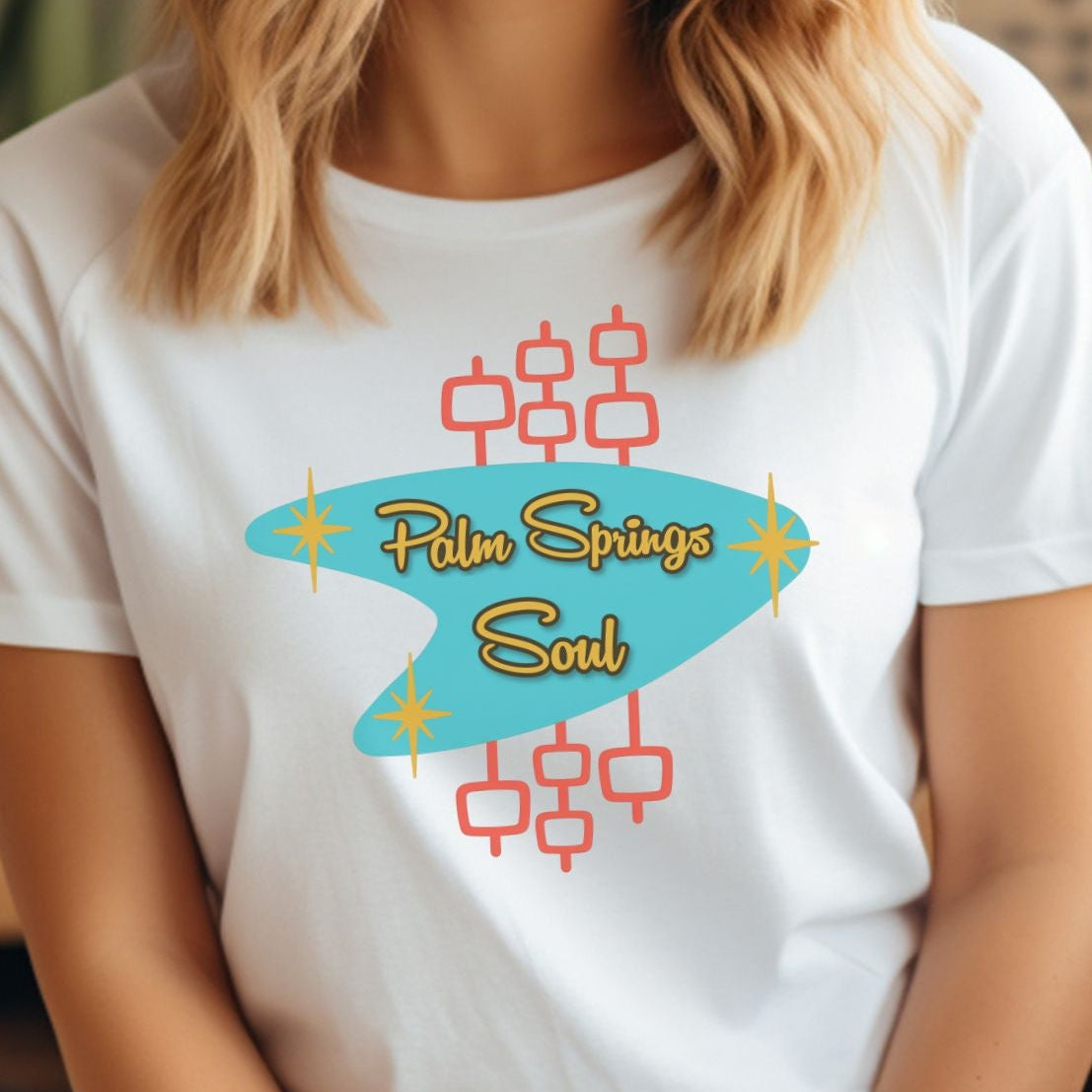 Person wearing a white t-shirt with colorful design and text, sitting indoors.