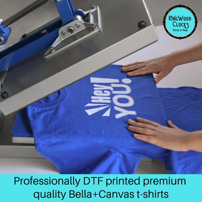 Blue t-shirt with a printed design being handled next to a screen printing machine, with 'Kingwood Clocks' branding.