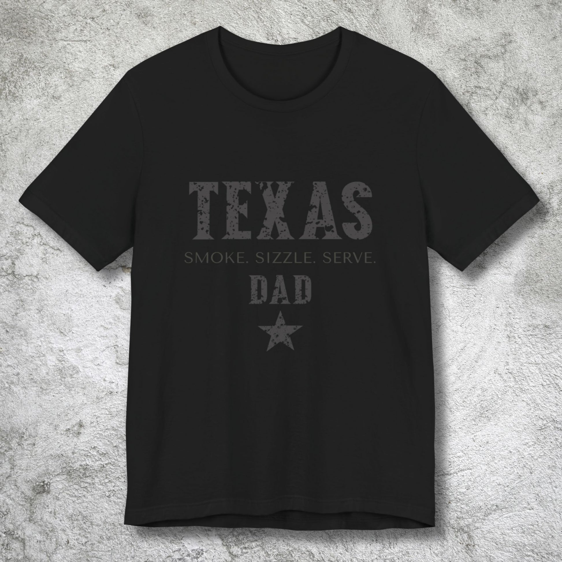 Black t-shirt with 'Texas Smoke, Sizzle, Serve Dad' text on a textured gray background
