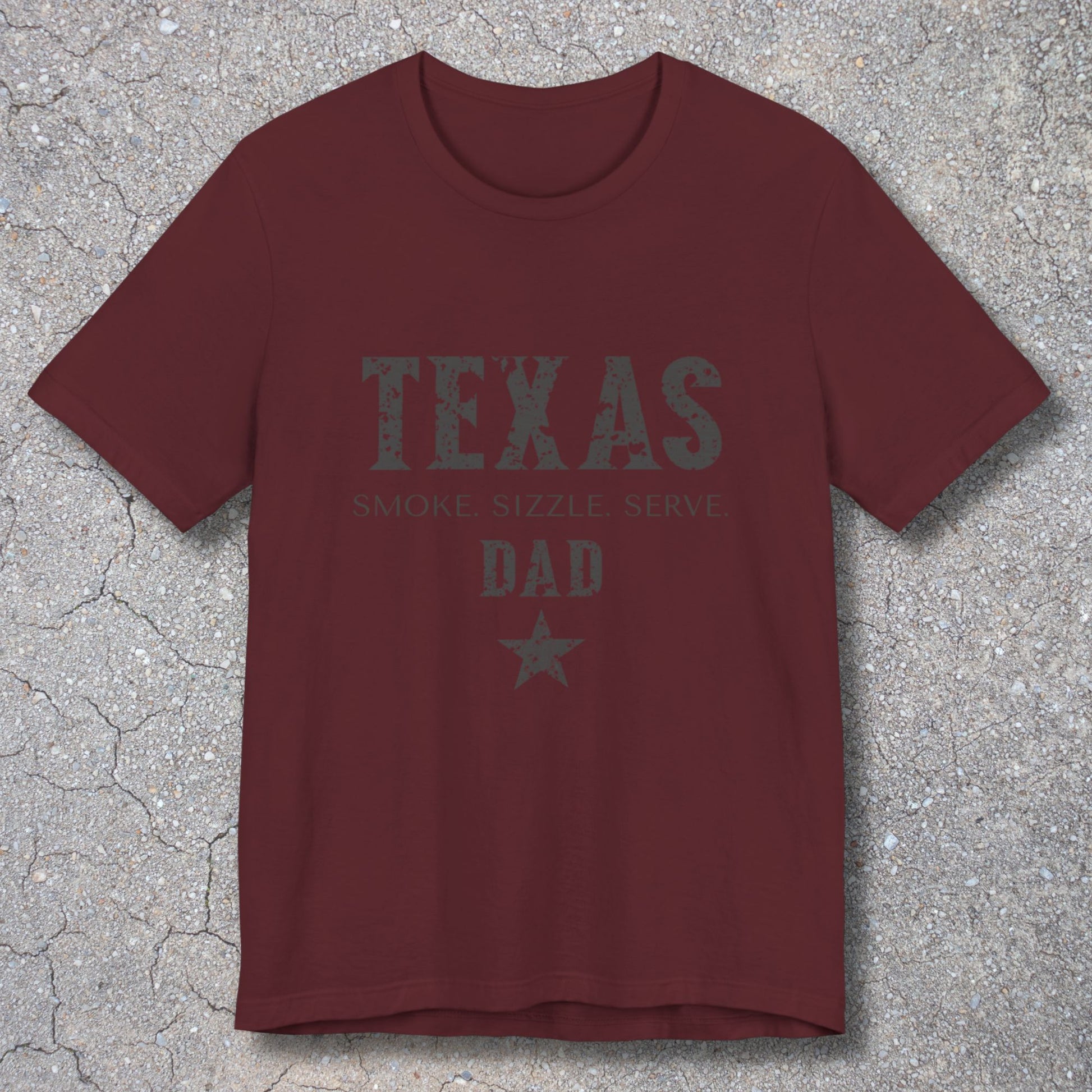Maroon t-shirt with 'Texas Smoke, Sizzle, Serve Dad' text on a textured surface