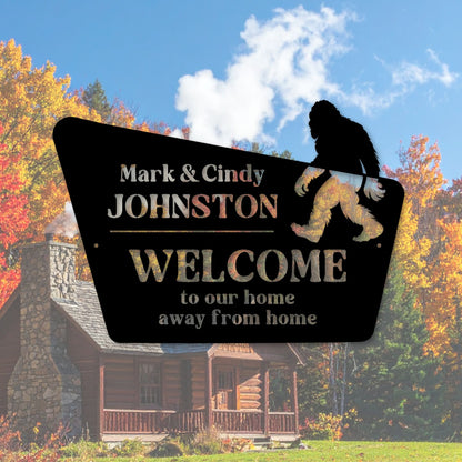 Bigfoot-themed welcome sign with personalized names in front of a log cabin during autumn.